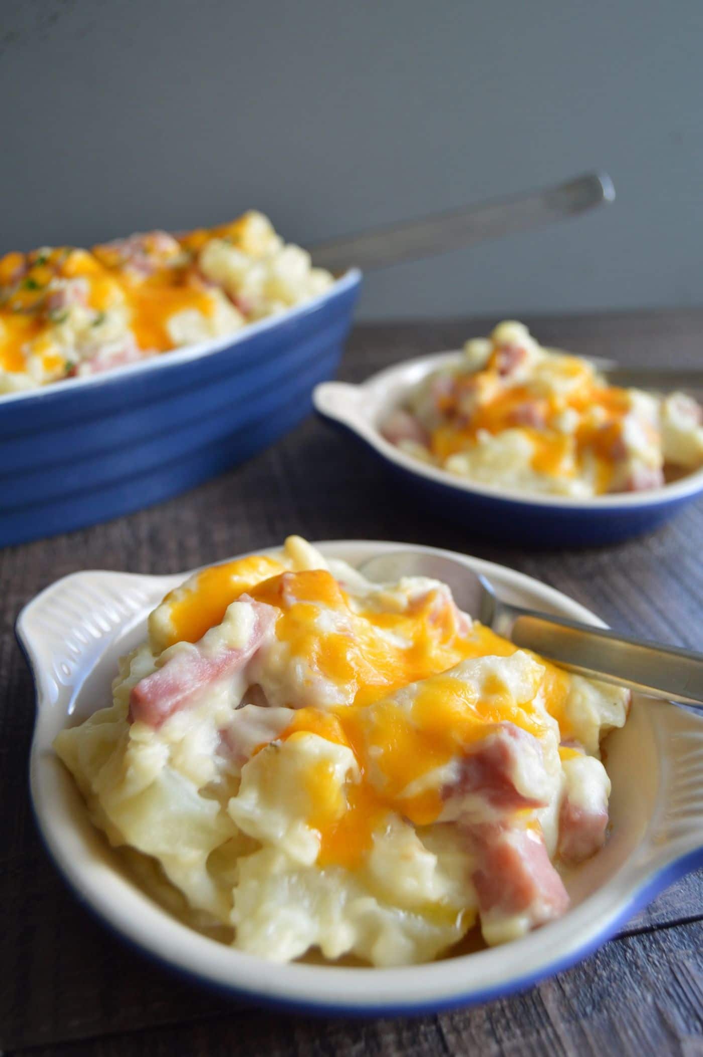 Gouda Ham and Potato Casserole in individual blue bowls with a large serving bowl with a spoon in it in the background on a table.