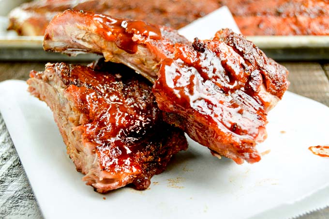 two pieces of ribs on a plate next to a tray with bbq sauce slathered on it