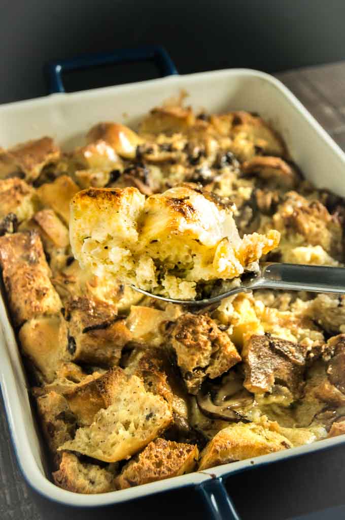  Baking dish with the fully cooked savory bread pudding with a spoon digging in and pulling out one small serving close up.