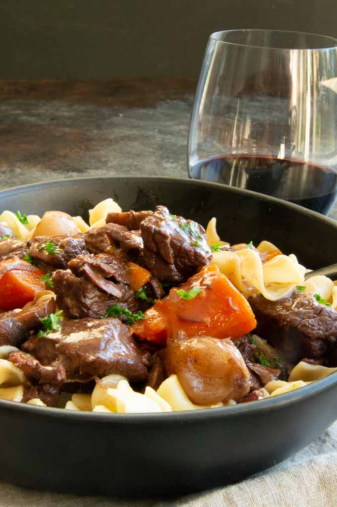A table setting of French beef stew in red wine sauce with a glass of wine next to it.
